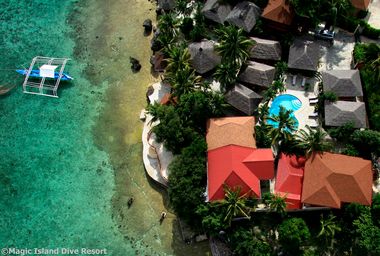 Aerial view of a beach resort with cottages, a pool, and a docked boat in clear turquoise water surrounded by lush greenery.