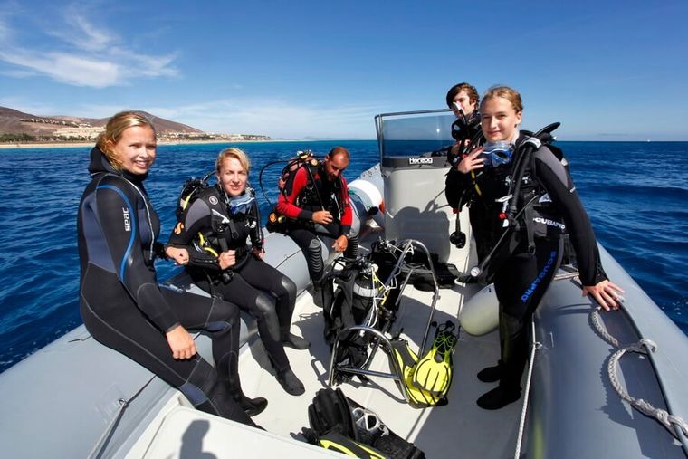 Duikers in wetsuits en uitrusting zitten op een boot op zee met helder blauw water en hemel.