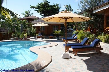 Outdoor pool area with blue lounge chairs, beige umbrellas, surrounded by tropical plants and cottages.
