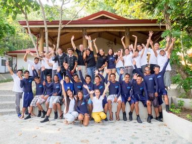 A large group of diverse people, wearing various casual outfits, smiling and raising their hands in front of a building.