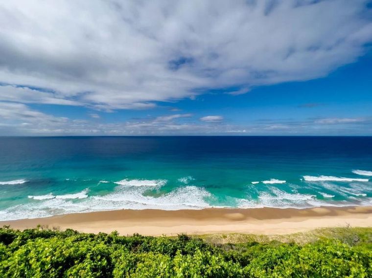 Sandy beach with turquoise ocean waves under a partly cloudy blue sky, bordered by green vegetation.