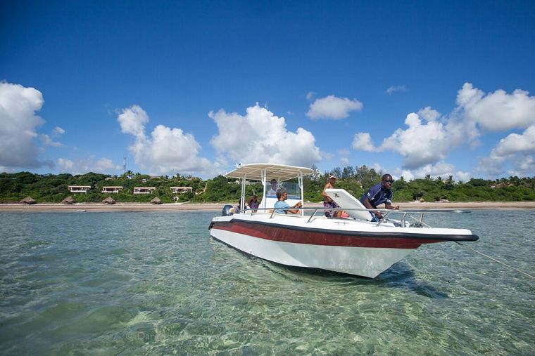 Boat on clear water near a sandy shore with green foliage and clouds in the blue sky.