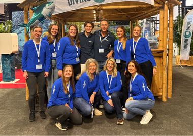 Group of people wearing blue shirts with name tags, posing in front of a diving-themed booth with a bamboo structure and decorative shark.